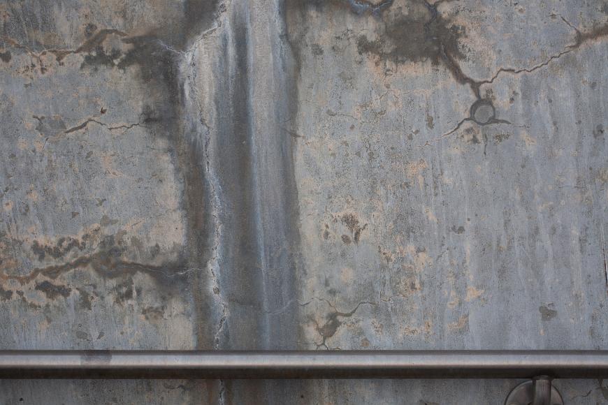 Detail Sullivan Station bus waiting area Somerville Massachusetts A metal handrail crosses horizontally at the bottom of the image, embedded in a concrete wall that has cracks, and dripping water stains in shades of grey and...