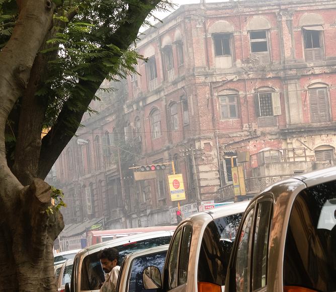 Old and new in downtown Kolkata Old brick building with weathered and in many places missing facade behind a row of newish looking cars with one driver waiting near the cars and a tree in the...
