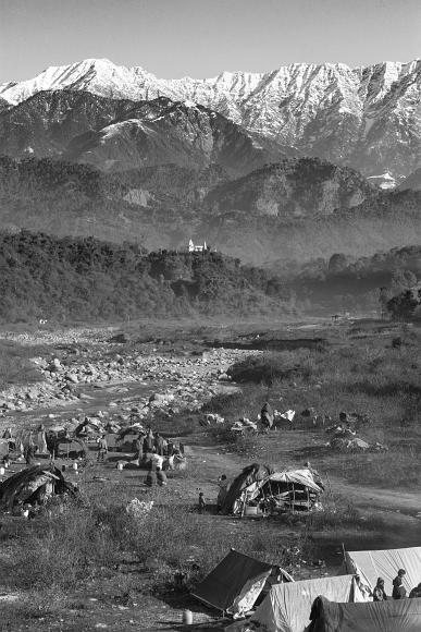 Peaks, Temple, River, Slum Snow covered peaks lie behind a temple positioned on a hill above a river that flows past a slum encampment in the foreground in the Himalayas near Dharamasala...