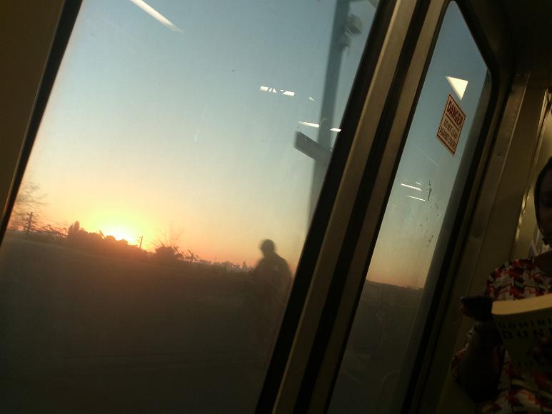 The view from BART Sun setting behing trees, loading cranes and the San Francisco city skyline as seen from inside a BART train at the West Oakland Station with a man on the...