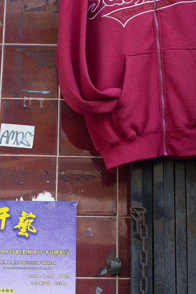 Sweatshirt and signs in Chinatown A maroon sweatshirt with the arms tucked into the pockets hangs above the open metal storefront grate while old tape and a poster in Chinese and English lie to...