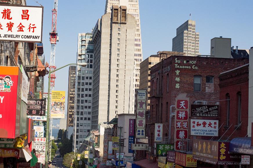 Chinatown street scene with downtown San Francisco in the background Many signs in Chinese and advertising Chineses businesses are visible on storefronts in Chinatown with the tall buildings on downtown and a sliver of the San...