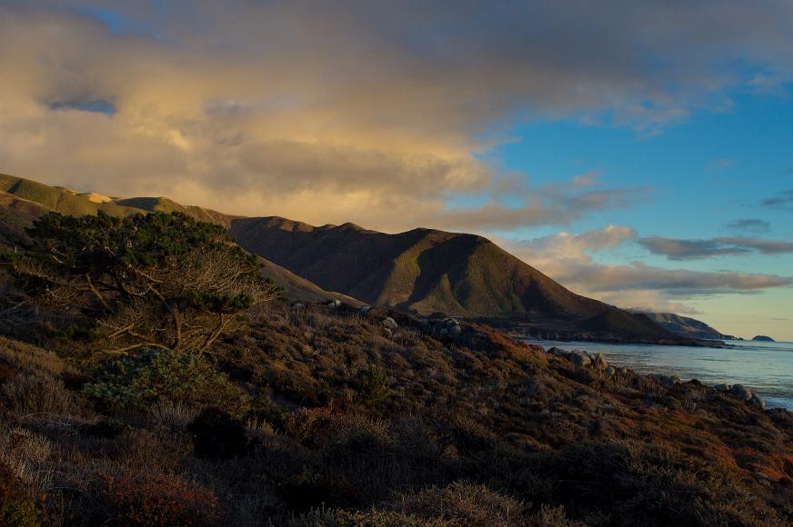 Central California Coast at sunset Mountains covered in scrub and pine trees glow in the last light above the Pacifc Ocean with yellow clouds above