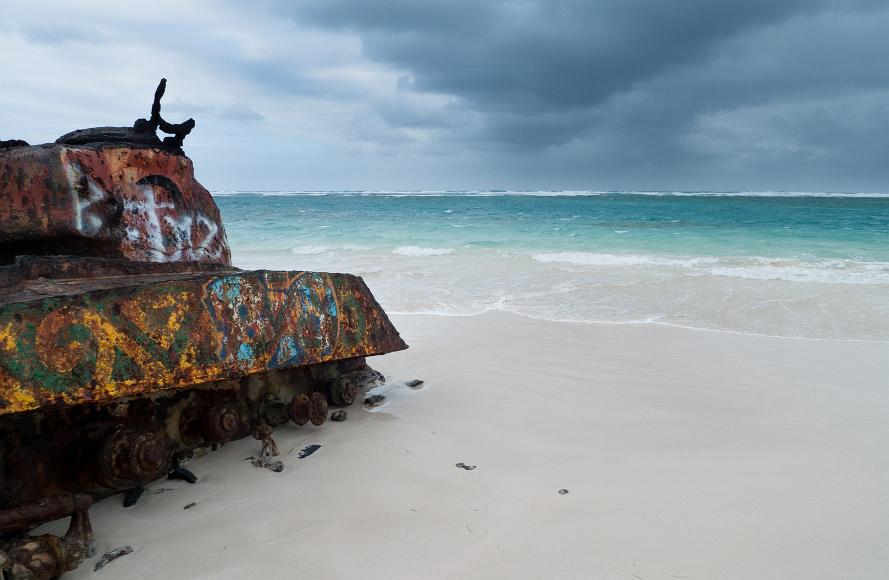 Military tank rests on a white sand beach in front of aquamarine waters and a stormy sky