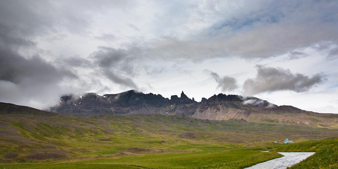 Jagged mountains and river valley in northeastern Iceland Jagged mountains and a mostly cloudy sky lie above a fertile green valley with one farmhouse and a rushing river in the foreground