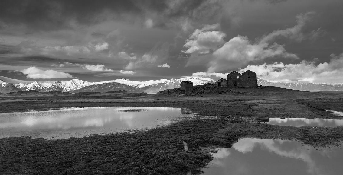 Hut against the light An abandoned house sits in front of snowcapped mountains and dramatic clouds in the sky, with the foreground comprising large still ponds refelcting the...