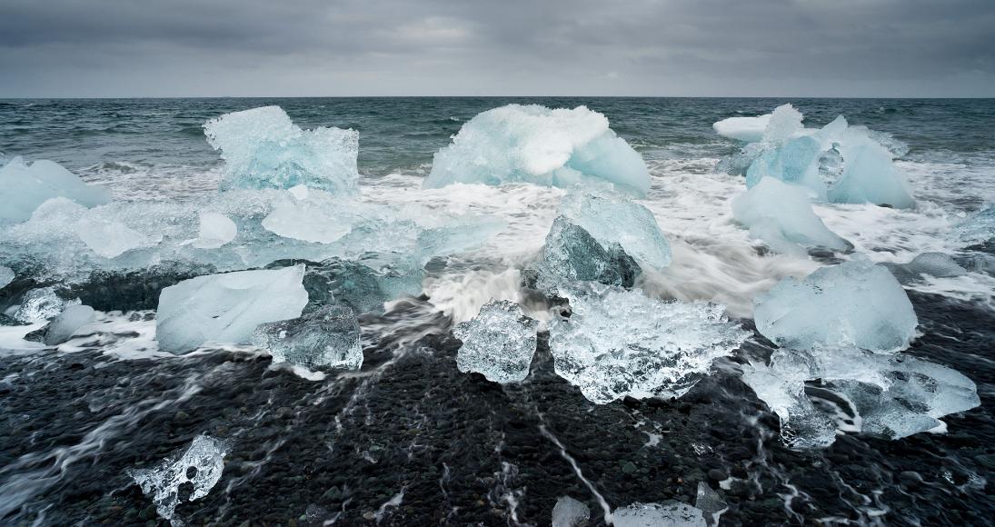 Ice on a black beach under dark clouds clear and translucent pieces of blue-ish pices of ice calved from a glacial toungue at Jokulsarlon, South Iceland
