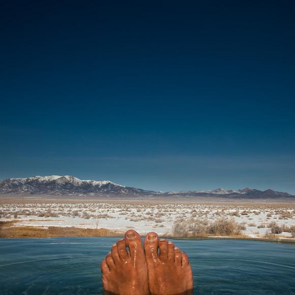 Happy feet belong in hot springs and hot springs belong in Nevada Two brown feet sticking out of a metal pool filled with hot water in front of a snowcovered desert valley with dark green and scnowcapped mountains in the...