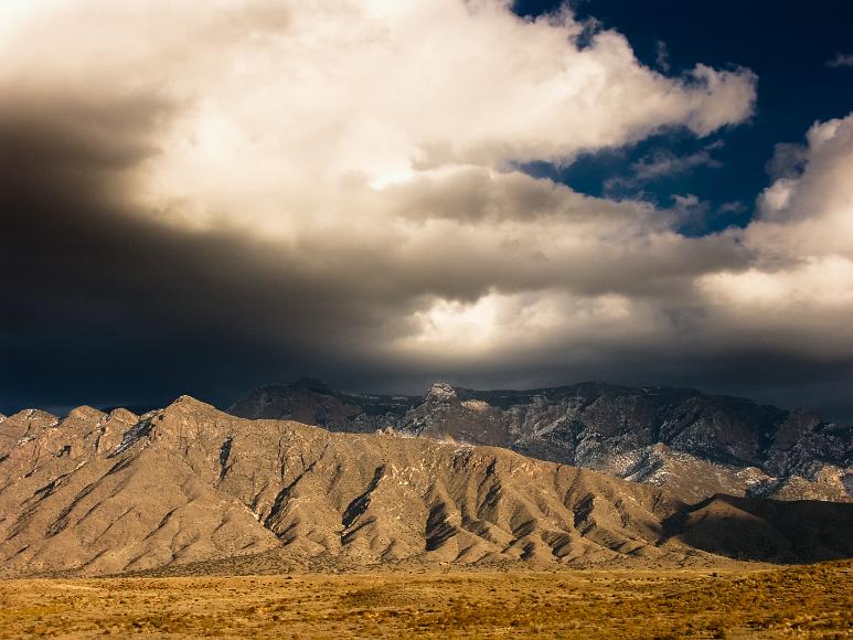 The Sandia Mountains at 85 miles an hour on I25, North of Albuquerque, New Mexico The scrubby flatlands (empty but for a lone power line) of the Pueblo of Sandia Village give way to deeply folded barren looking hills with the Sandia massif...