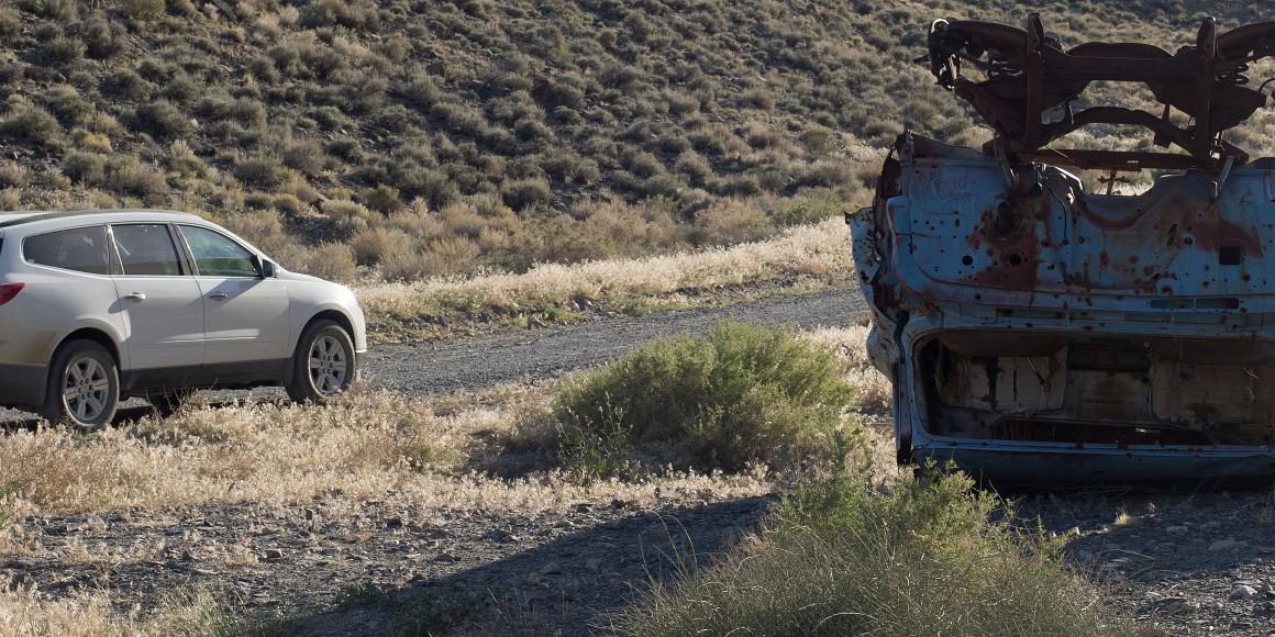 New car, upside down shot car A Chevy Traverse pussy SUV is on a road by a much more hardcore old 50s cars that is light blue and completely riddled with bullets