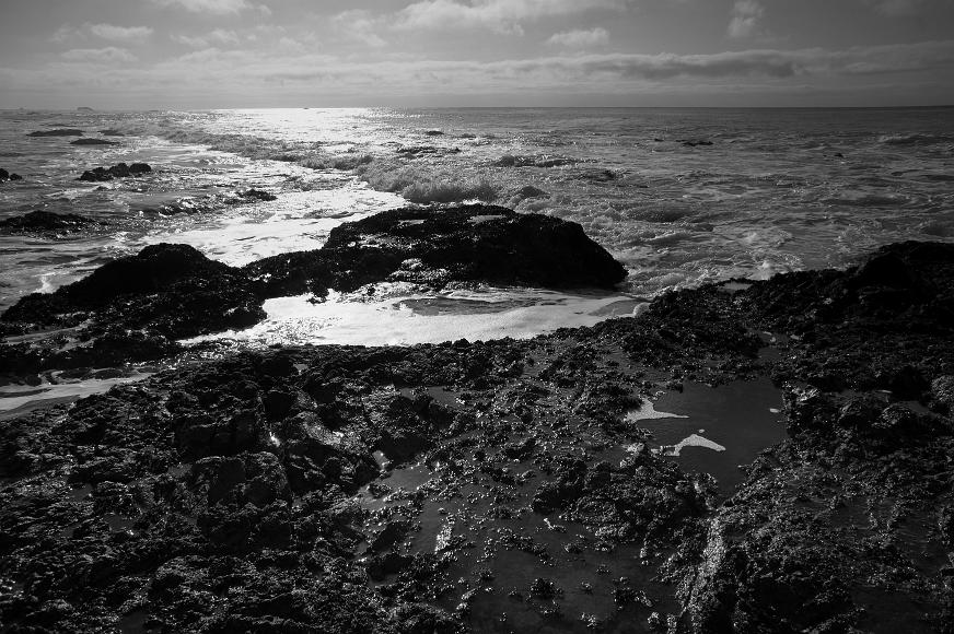 Water crashing on a rocky shore Late afternoon sunshine from a partly cloudy sky shines against waves crashing against a very rocky volcanic shore and settling into foamy tidepools near...