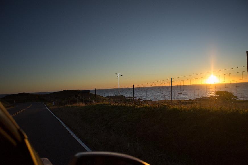 Sunset from highway one Yellow sun setting over the blue pacific ocean with trees and grass on the coast as seen through a wire cattle fence from the passenger side of a car travelling...