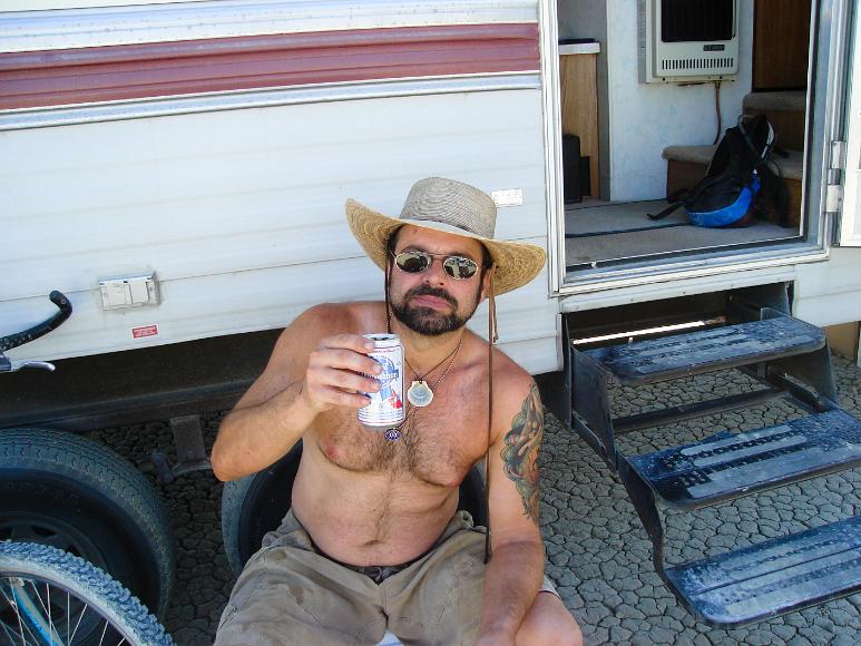Ron lifts a cold one A man holds a open PBR towards the viewer as he sits on dry lakebed in front of the open door of a recreational vehicle