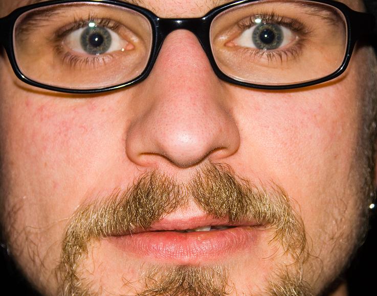 Phils face closeup of a face with some facial hair and black plastic framed glasses lit by a direct flash