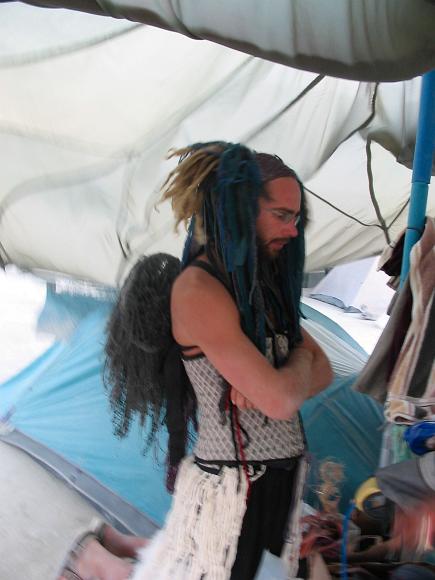 Chris in full Red Ant fashion show regalia A very tan man with a curly blond dreadlocks with fabric intertwined wears a corset and rope skirt underneath a parachute