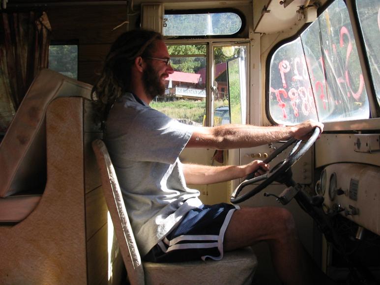 Crhis behind the wheel of the bus A man with dreadlocks sits smiling at the wheel of a very old bus that has for sale written on the window alongside a phone number