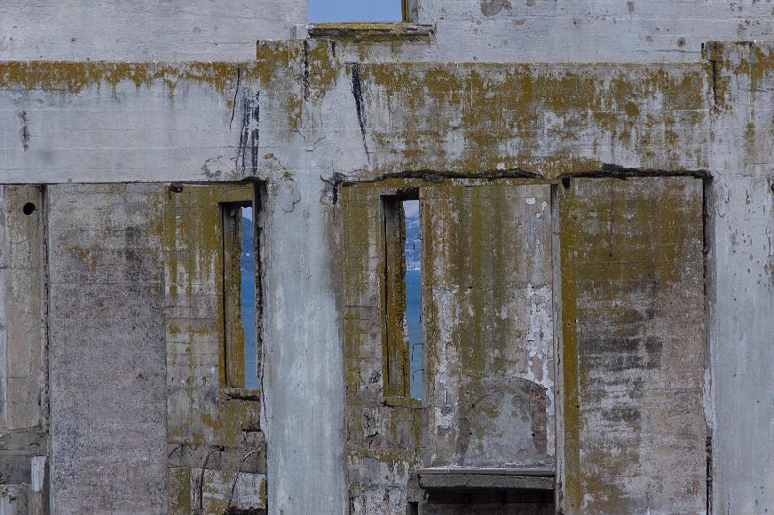 Looking out from jail Gray concrete wall with green moss growing on it punctuated by small slit windows allowing a view to the San Francisco Bay beyond and sailboats and Marin County...