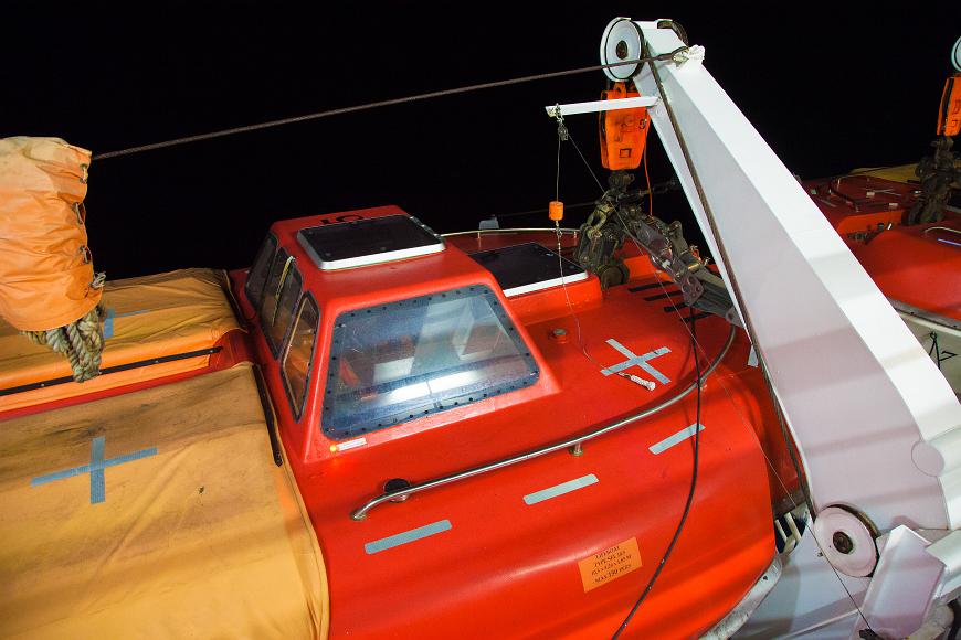 Modern lifeboat at night on a Carnival Cruise ship Modern fully enclosed lifeboat painted bright red with solar panels on top against an inky black nighttime sea, suspended from the side of a Carnival Cruise...